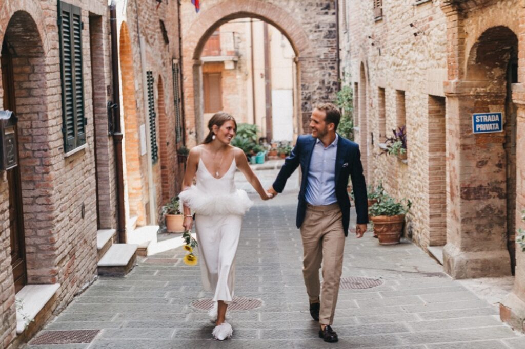 Ein lächelndes Paar in halb-formeller Kleidung geht Hand in Hand eine bezaubernde, schmale, kopfsteingepflasterte Straße hinunter, die an eine Villa Cetinale-Hochzeit erinnert, und hält Blumen und genießt die Gesellschaft des anderen.