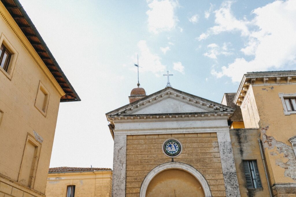 Historische gelbe Steingebäude mit Ziegeldächern umgeben ein zentrales Gebäude mit einem dreieckigen Giebel, einem runden Emblem und einem Kreuz auf der Spitze - eine elegante Kulisse, perfekt für eine Villa Cetinale Hochzeit unter dem hellen, teilweise bewölkten Himmel.
