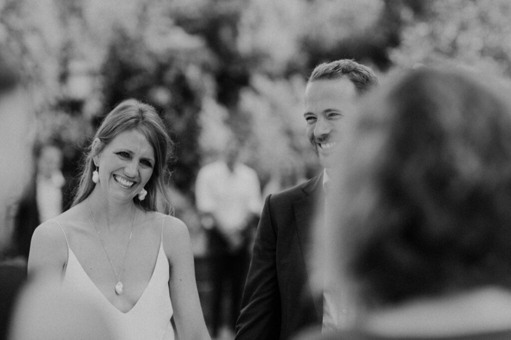 Ein Schwarz-Weiß-Foto eines lächelnden Paares im Freien, möglicherweise bei einer Hochzeit in der Villa Cetinale. Die Frau trägt ein helles Kleid mit Ohrringen, der Mann einen Anzug. Beide wirken glücklich, umgeben von unscharfen Gästen und Grünzeug.