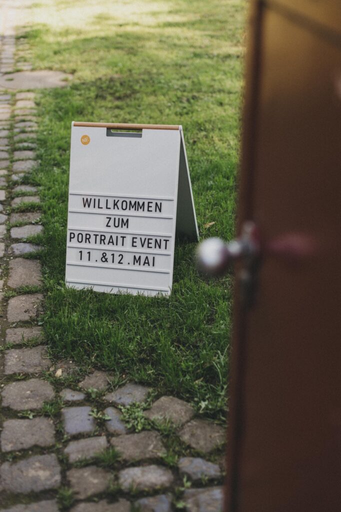 Auf einem weißen Schild im Gras steht "Willkommen zum Portrait Event 11. & 12. Mai. Perfekt für Kids Fotograf Sessions, mit einem Teil einer Tür und einem Türknauf, der am rechten Rand des Bildes zu sehen ist.