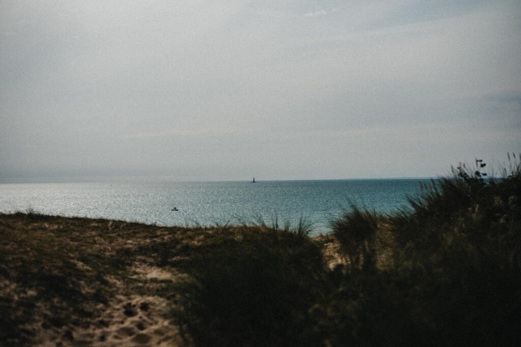 Ein Sandstrand mit grasbewachsenen Dünen im Vordergrund blickt auf ein ruhiges, blaues Meer unter einem dunstigen Himmel. Aufgenommen von einem talentierten Fotografen Köln, sind ein kleines Boot und ein entfernter Leuchtturm am Horizont zu sehen.