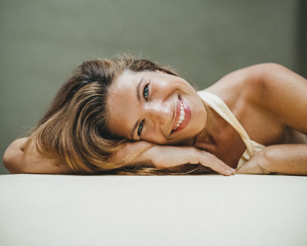 Eine Frau mit langen braunen Haaren liegt auf der Seite, stützt ihren Kopf und ihren Arm auf eine weiße Fläche und lächelt warm in die Kamera vor einem weichen, neutralen Hintergrund - aufgenommen von einem Familienfotografen in Köln.