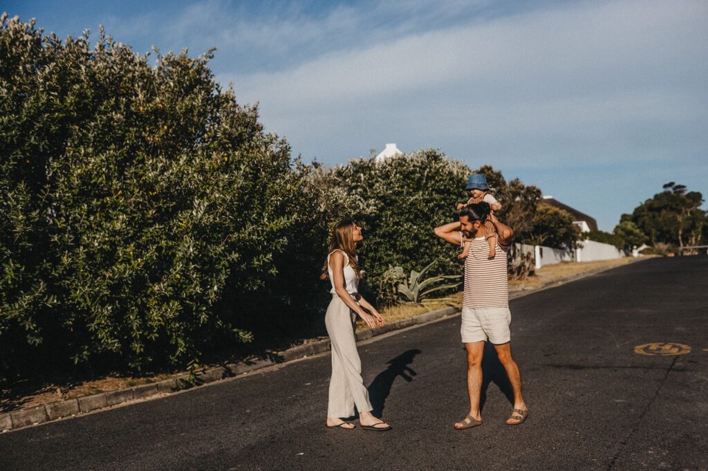 Eine Frau und ein Mann stehen in einer ruhigen Vorstadtstraße, der Mann trägt ein kleines Kind auf seinen Schultern. Aufgenommen von einem Fotografen Köln, genießen sie einen sonnigen Tag mit Bäumen und Häusern im Hintergrund.