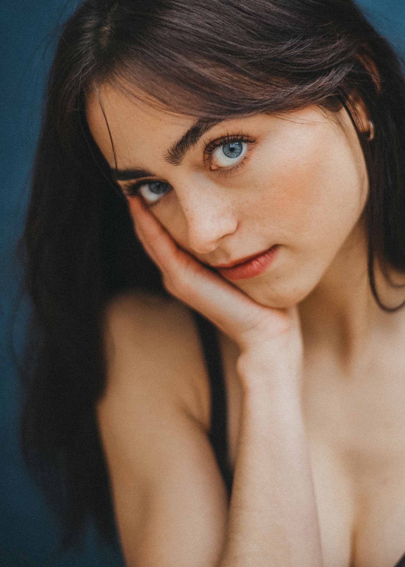 Eine junge Frau mit langen dunklen Haaren und blauen Augen stützt ihr Gesicht auf ihre Hand und blickt nachdenklich in die Kamera vor einem dunkelblauen Hintergrund - aufgenommen von einer erfahrenen Fotografin in einem renommierten Fotostudio Köln.