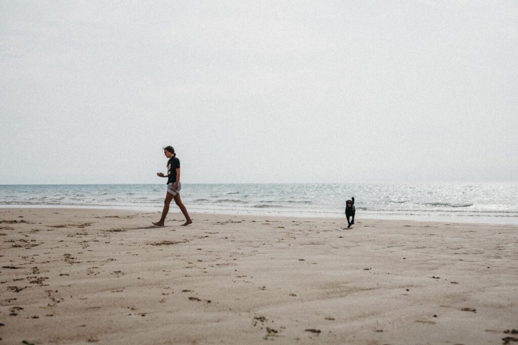 Eine Person läuft barfuß an einem Sandstrand in der Nähe der Wasserkante entlang, dicht gefolgt von einem kleinen schwarzen Hund. Das ruhige Meer und der bedeckte Himmel, aufgenommen von einem talentierten Werbefotografen, schaffen eine heitere Stimmung, die an das Fotostudio Köln erinnert.