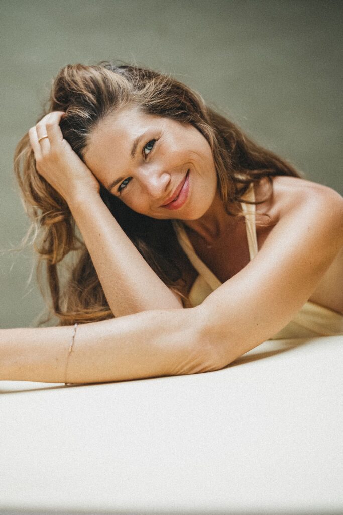 Eine Frau mit langen braunen Haaren und einem hellgelben Oberteil stützt ihren Arm auf einer weißen Fläche ab und lächelt warm in die Kamera, während sie ihre Hand in den Haaren hält - ein perfekter Moment, eingefangen von einem talentierten Familienfotografen. Der Hintergrund ist sanft verschwommen.