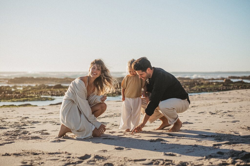Eine Frau, ein Mann und ein Kind lächeln und spielen zusammen an einem Sandstrand in der Nähe des Wassers unter einem klaren blauen Himmel und genießen einen sonnigen Tag - eingefangen von einem erfahrenen Kinderfotografen.