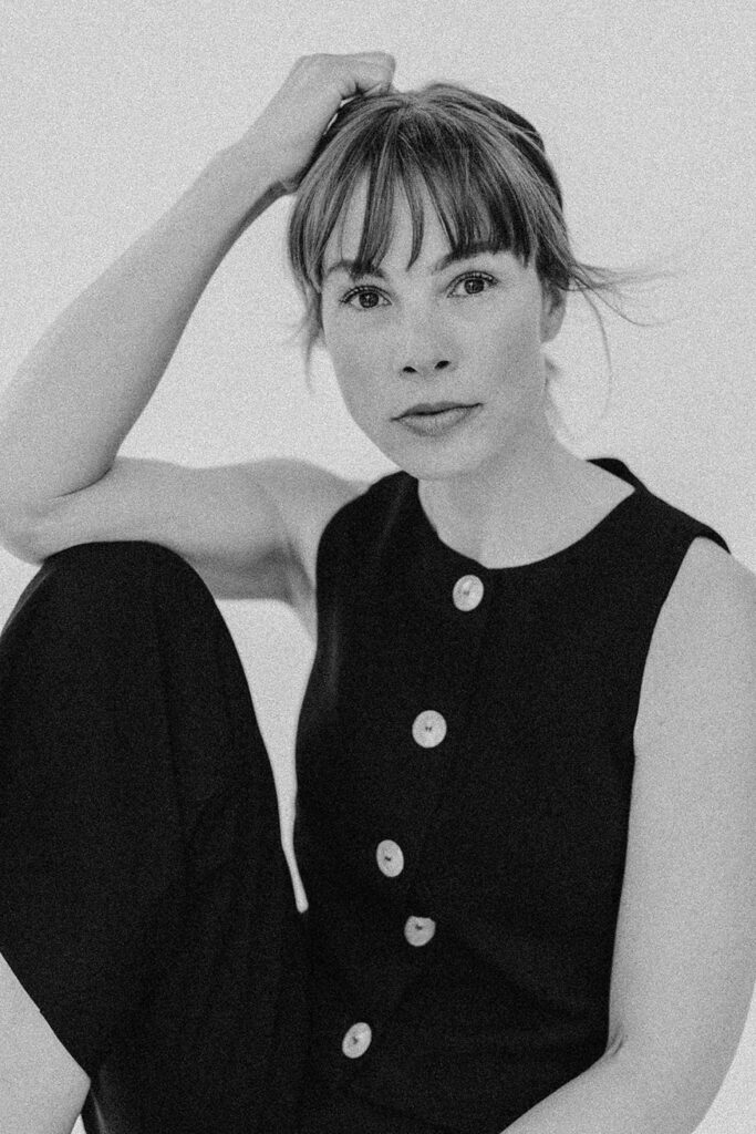 Schwarz-Weiß-Foto einer Frau, die mit dem rechten Arm auf dem Kopf sitzt und ein ärmelloses schwarzes Oberteil mit großen Knöpfen trägt - aufgenommen von einer Fotografin in Köln, blickt sie ruhig in die Kamera.