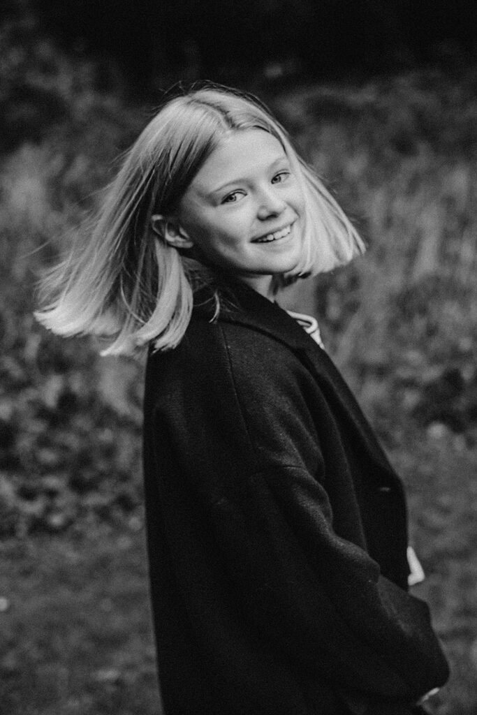 Schwarz-Weiß-Foto einer jungen Person mit kinnlangen hellen Haaren, lächelnd in einem dunklen Mantel, die sich der Kamera im Freien zuwendet, mit unscharfem Gras im Hintergrund - aufgenommen von Familien Fotograf im Fotostudio Köln.