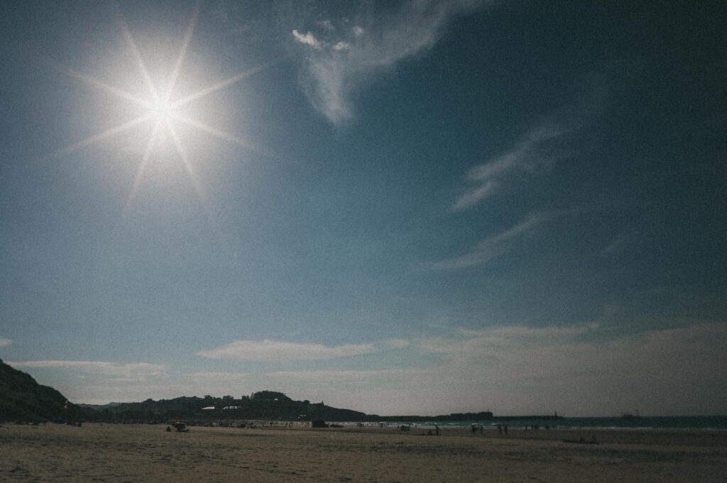 Die Sonne scheint hell am meist wolkenlosen Himmel über einem Sandstrand, und in der Ferne sind ein paar Menschen in der Nähe des Ufers zu sehen - ein ideales Motiv für jeden Kinderfotografen oder Werbefotografen, der natürliche Momente vor dem Hintergrund niedriger Hügel festhalten möchte.