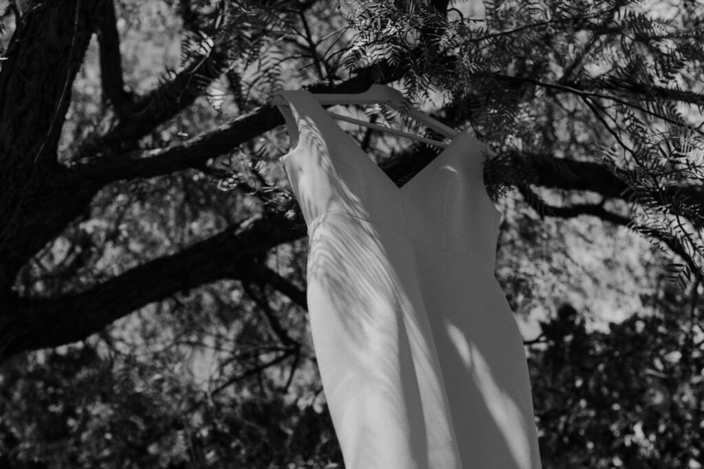 Ein ärmelloses weißes Kleid hängt von einem Baumzweig herab, wobei das Sonnenlicht und die Schatten der Blätter Muster auf dem Stoff erzeugen. Das in Schwarz-Weiß aufgenommene Foto strahlt eine sanfte, elegante Atmosphäre aus - die perfekte Inspiration für eine verträumte Hochzeit in Sizilien.