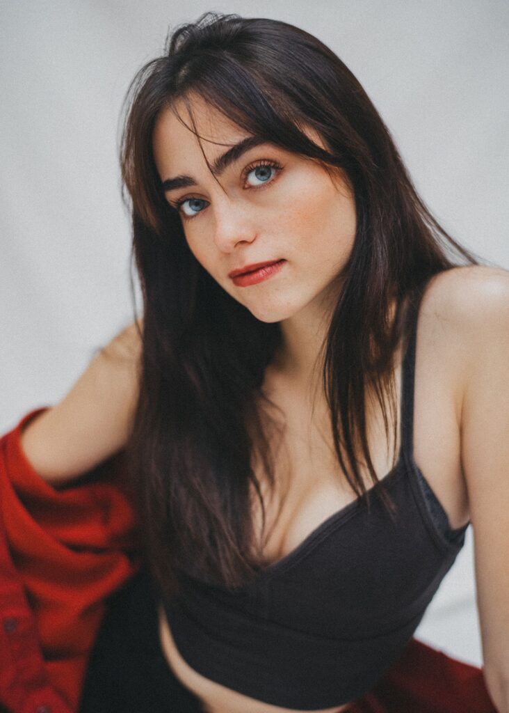 Eine junge Frau mit langen dunklen Haaren und blauen Augen blickt in die Kamera. Die von einem Familienfotografen aufgenommene Frau trägt ein schwarzes Crop-Top und ein rotes Hemd, das vor einem hellen Hintergrund über ihre Schulter fällt.