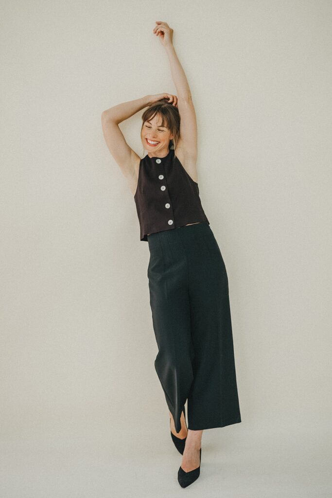 Eine Frau in einem ärmellosen schwarzen Oberteil mit großen Knöpfen und einer schwarzen Hose mit weitem Bein posiert spielerisch vor einem schlichten, hellen Hintergrund. Sie lächelt und hebt einen Arm über den Kopf - aufgenommen von einer talentierten Fotografin im Fotostudio Köln.