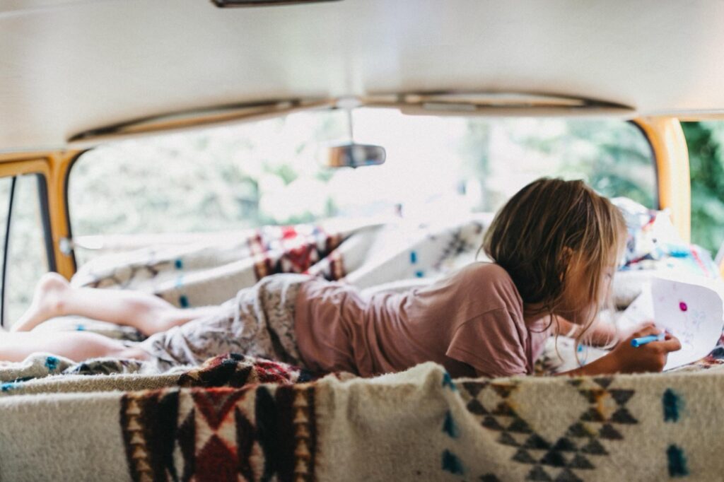 Ein Kind liegt auf dem Bauch in einem Wohnmobil und malt auf Papier. Gemusterte Decken bedecken das Bett und Sonnenlicht strömt durch die Fenster und schafft eine gemütliche Atmosphäre - perfekt, um von einem Kids Fotograf oder Fotostudio Köln festgehalten zu werden.