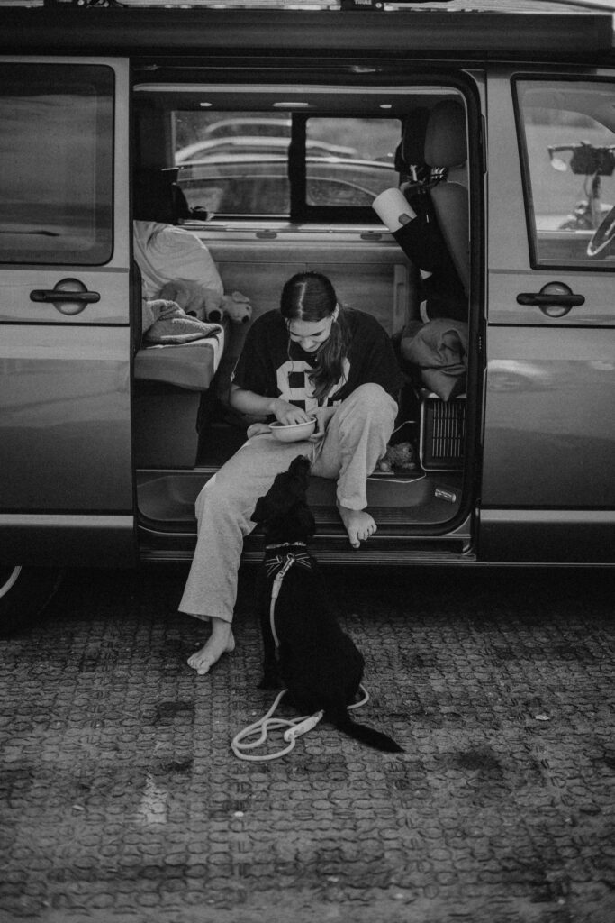 Eine barfüßige Frau sitzt in der offenen Schiebetür eines Lieferwagens und lächelt auf ihren schwarzen Hund an der Leine herab. Umgeben von ihren Habseligkeiten strahlt sie Wärme aus - ein ehrlicher Moment, eingefangen von einem talentierten Familien Fotograf Köln.