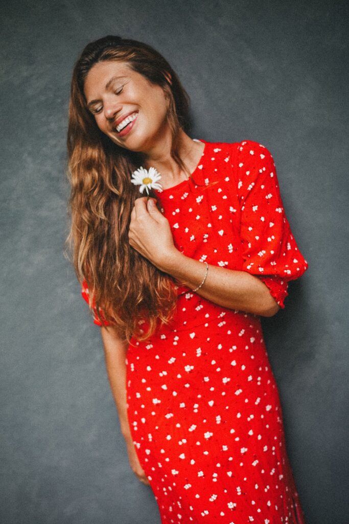Eine Frau in einem roten Kleid mit weißen Blumenmustern lächelt mit geschlossenen Augen und hält ein Gänseblümchen an ihr Gesicht. Sie hat langes braunes Haar und steht vor einem grauen, strukturierten Hintergrund, der von einem Familienfotografen in Köln aufgenommen wurde.