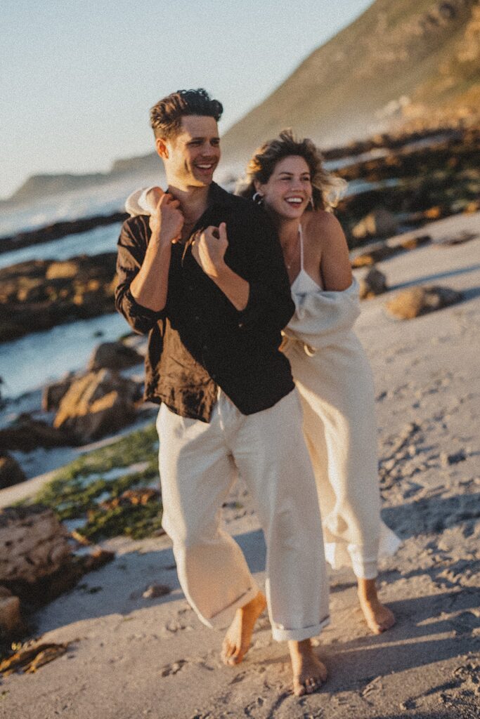 Ein lächelndes Paar geht bei Sonnenuntergang barfuß an einem felsigen Strand spazieren. Der Mann trägt ein schwarzes Hemd und eine helle Hose, die Frau in Weiß hält seinen Arm, aufgenommen von einer erfahrenen Fotografin. Wellen und Hügel zieren den Hintergrund - Momente vom Fotostudio Köln.