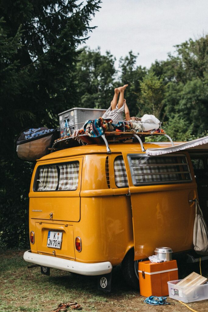 Ein gelber Oldtimer-Van, der von einem Werbefotografen aufgenommen wurde, ist im Freien geparkt und mit Gepäck und Campingausrüstung beladen. Die Beine einer Person ragen aus dem Dach, umgeben von Bäumen und verstreutem Campingzubehör.