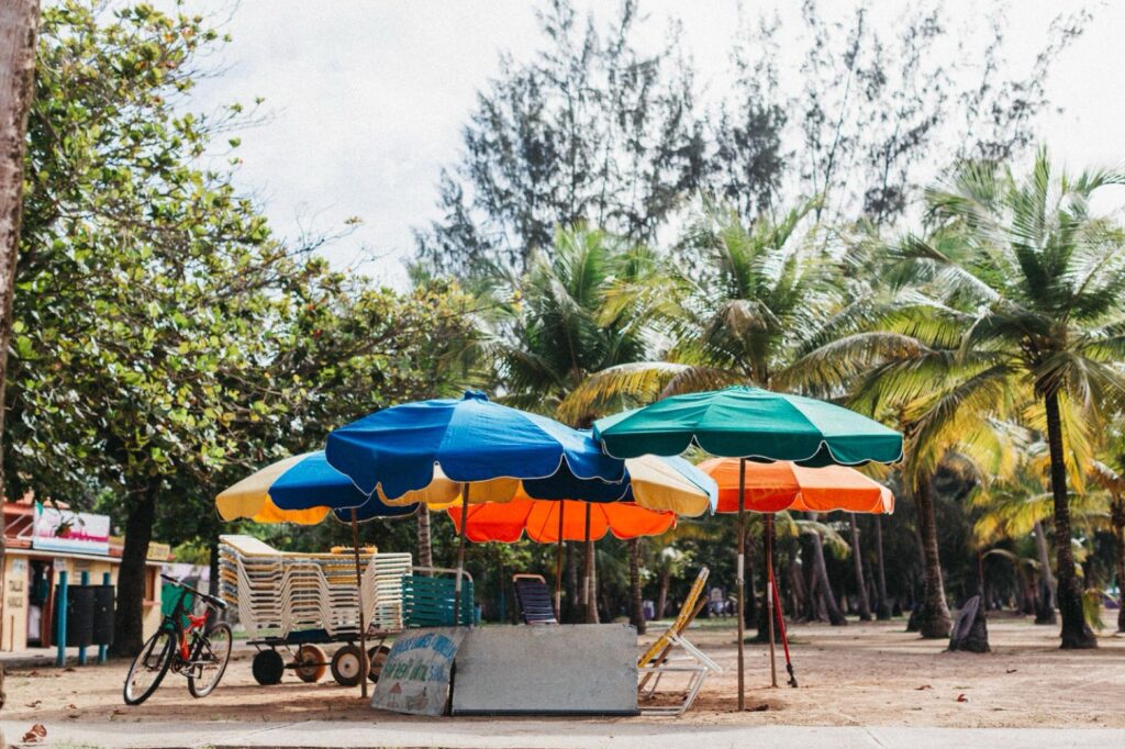 Bunte Sonnenschirme und gestapelte Liegestühle stehen auf sandigem Grund, dahinter Palmen, ein Fahrrad lehnt in der Nähe. Diese ruhige, tropische Szene wurde von einem Werbefotografen aufgenommen und erinnert an ein Fotoshooting im Fotostudio Köln am Strand.