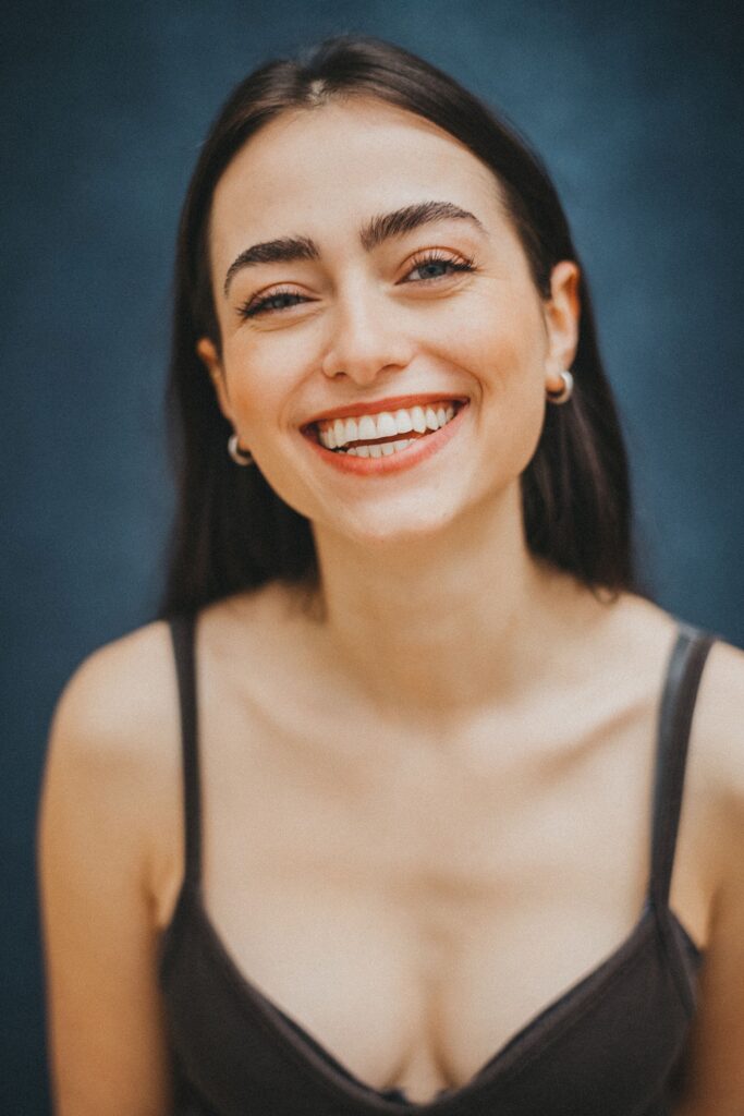 Eine junge Frau mit langen braunen Haaren lächelt strahlend in die Kamera. Aufgenommen von einem talentierten Fotografen Köln, trägt sie ein schwarzes ärmelloses Oberteil und kleine Reifohrringe. Der Hintergrund ist dunkelblau und sanft verschwommen.