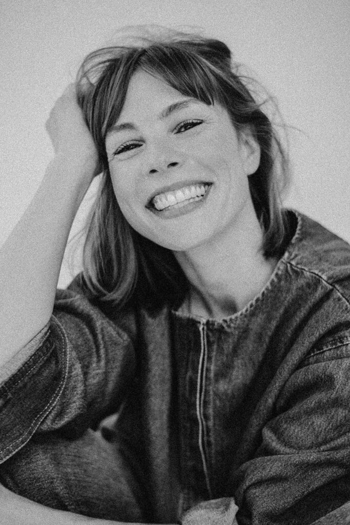 Schwarz-Weiß-Foto einer Frau mit schulterlangem Haar, die eine Jeansjacke trägt und breit in die Kamera lächelt. Aufgenommen von einem Familien Fotograf im Fotostudio Köln, stützt sie ihren Kopf auf ihre Hand und wirkt fröhlich und entspannt.