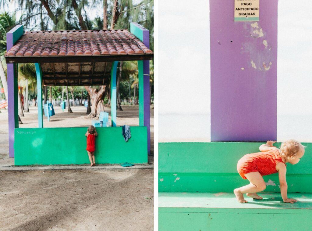 Ein Kleinkind in einem roten Outfit spielt in der Nähe eines bunten Unterstandes mit grünen und lila Wänden in einem Park am Strand, aufgenommen von einem Werbefotografen. Das Kind erkundet die niedrige Mauer, während im Hintergrund Bäume und Sand zu sehen sind.