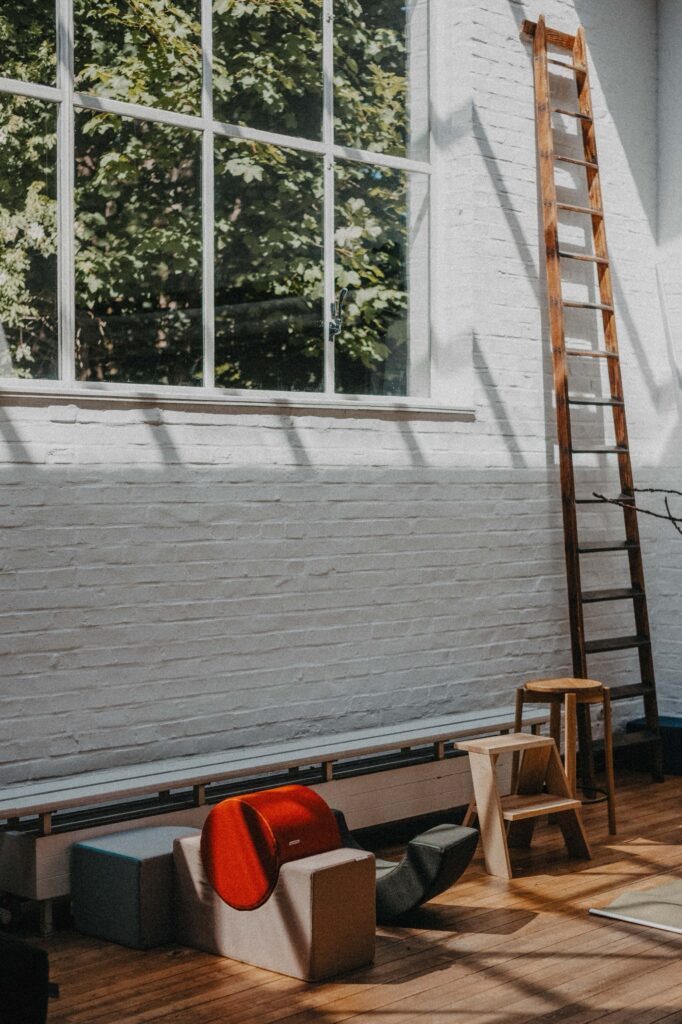 Ein heller Raum mit großen Fenstern, einer hölzernen Leiter, die an eine weiße Backsteinwand gelehnt ist, und gemütlichen Hockern und Kissen auf dem Boden - ein einladender Raum, der sich perfekt für eine Familien Fotograf Session oder kreative Shootings von Fotograf Köln eignet. Das Sonnenlicht wirft sanfte Schatten ins Innere.