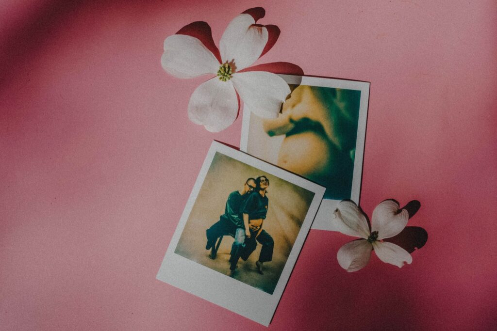 Zwei Polaroidfotos und weiße Hartriegelblüten auf rosa Hintergrund. Aufgenommen von einem Kinderfotografen aus einem Fotostudio in Köln, zeigt das eine Foto zwei eng beieinander sitzende Personen, das andere ist ein unscharfes, abstraktes Bild.