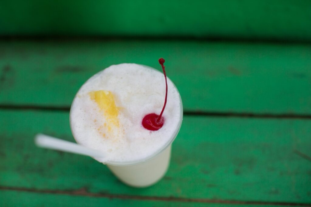 Ein cremiges Getränk in einem durchsichtigen Plastikbecher mit weißem Strohhalm, gekrönt von schäumendem Schaum, einer Kirsche und einem Ananasstück, wird von einem Familienfotografen kunstvoll in Szene gesetzt und auf einer grünen Holzfläche platziert.