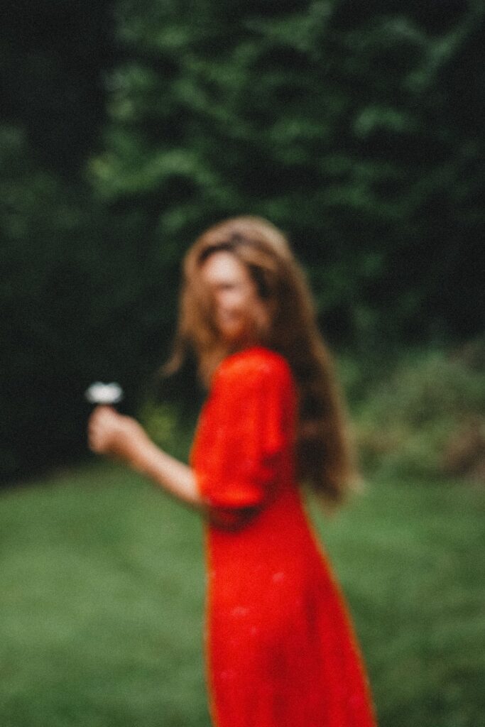 Eine Person in einem roten Kleid steht draußen im Gras und hält eine weiße Blume; das Bild ist sehr unscharf, mit grünem Laub im Hintergrund - perfekt für ein verträumtes Kinderfotoshooting oder eine kreative Session im Fotostudio Köln.