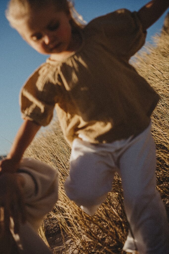 Ein kleines Kind in einem hellbraunen Hemd und einer weißen Hose läuft durch hohes, goldenes Gras unter einem klaren blauen Himmel. Das von einem Werbefotografen aufgenommene, weich fokussierte Bild vom Fotostudio Köln vermittelt Bewegung und Wärme.