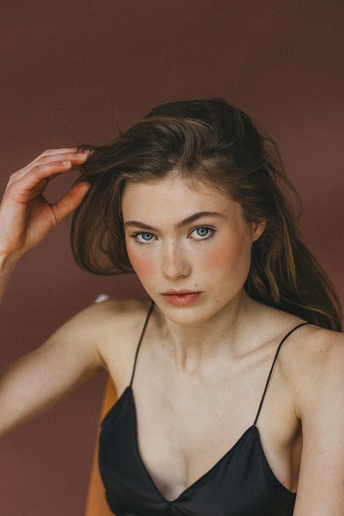 Eine junge Frau mit heller Haut und blauen Augen, die ein schwarzes Spaghettiträger-Top trägt, blickt direkt in die Kamera. Sie berührt mit einer Hand ihr Haar vor einem schlichten braunen Hintergrund in einem Fotostudio Köln.