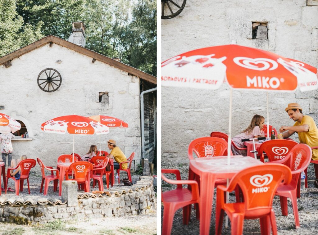 Zwei Fotos eines Familienfotografen zeigen ein Straßencafé mit roten "Miko"-Regenschirmen. Ein Mann und ein Kind genießen eine Mahlzeit, während eine Frau in der Nähe steht, alles neben einem rustikalen, begrünten Steingebäude im Hintergrund.