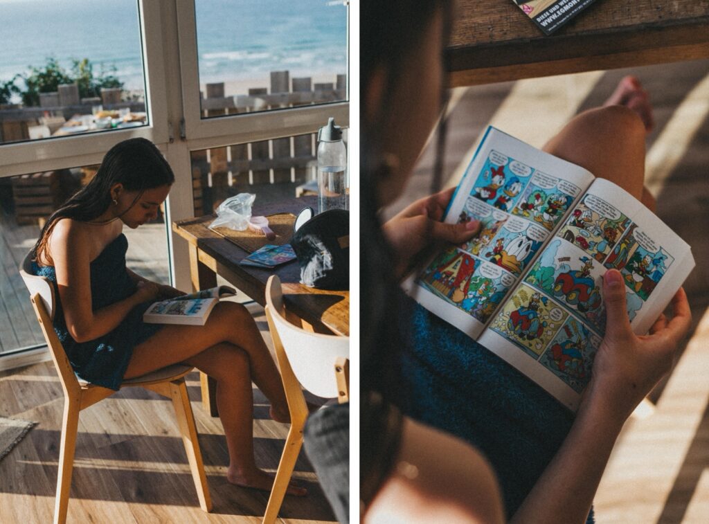 Ein Mädchen in einem blauen Handtuch sitzt an einem sonnenbeschienenen Fenster und liest einen bunten Comic; eine Nahaufnahme zeigt ihre Hände, die den aufgeschlagenen Comic halten, mit einem Tisch und Meerblick im Hintergrund - aufgenommen von einem Familien Fotograf in Köln.