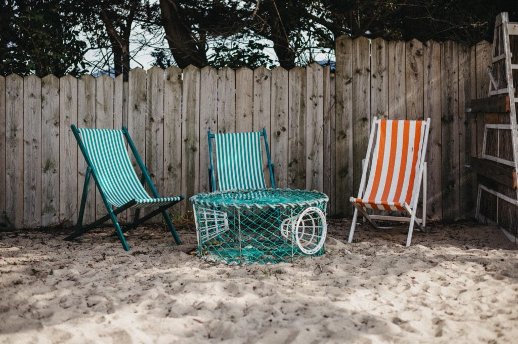 Drei gestreifte Liegestühle - zwei blau-weiße und ein orange-weißer - stehen um einen runden, grünen Drahttisch auf sandigem Boden. Im Hintergrund befinden sich ein Holzzaun und Bäume, die eine entspannte Szene schaffen, die perfekt für einen Familien Fotograf oder ein Fotostudio Köln ist.