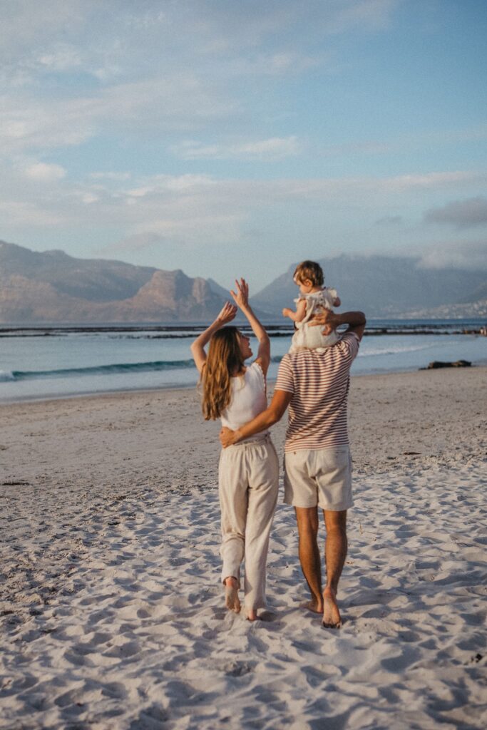 Eine dreiköpfige Familie spaziert barfuß über einen Sandstrand in Richtung Meer und verkörpert damit einen perfekten Moment für einen Familienfotografen. Die Frau hat ihren Arm um den Mann gelegt, der ihr Kind auf den Schultern trägt. Berge und blauer Himmel vervollständigen die heitere Kulisse.