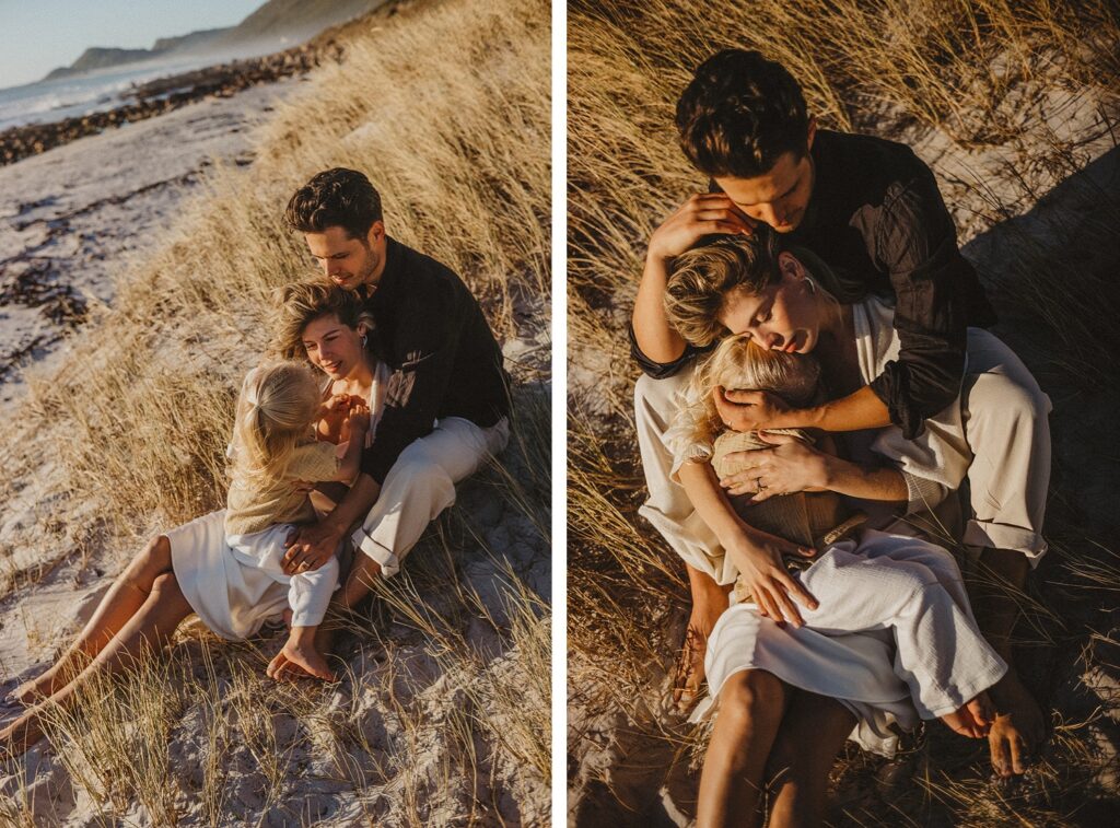 Ein Paar sitzt eng beieinander an einem Sandstrand inmitten von hohem Gras, umarmt sich und lächelt, während warmes, goldenes Sonnenlicht ein sanftes Licht wirft - eingefangen von Familien Fotograf für zeitlose Erinnerungen.