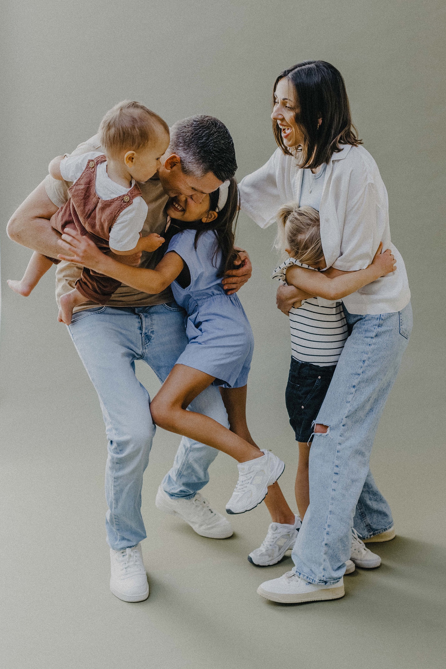 Eine fröhliche vierköpfige Familie, die von einem Familienfotografen aufgenommen wurde, umarmt und lacht zusammen. Ein Erwachsener hält ein Kleinkind im Knien, die anderen umarmen sich, alle lächeln in legerer Kleidung vor einem neutralen Hintergrund.