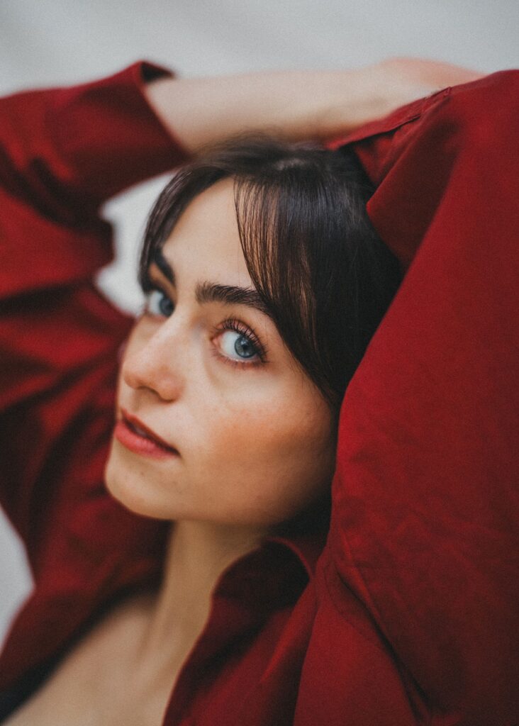 Eine Person mit heller Haut, blauen Augen und dunklen Haaren posiert mit erhobenen Armen in einem roten Hemd. Als Werbefotograf in Köln hebt der unscharfe Hintergrund ihr Gesicht und den intensiven Blick in die Kamera hervor.