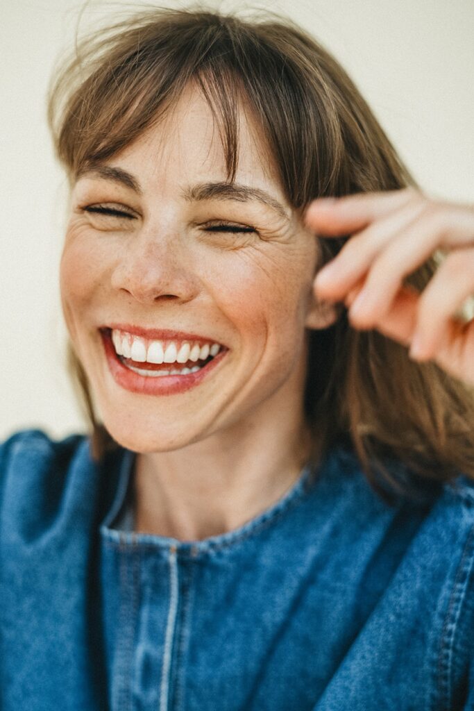 Eine Frau mit hellbraunem Haar und Pony, die ein Jeansoberteil trägt, lächelt fröhlich mit geschlossenen Augen. Die von einem Werbefotografen aufgenommene Frau lacht und hebt eine Hand neben ihrem Gesicht vor einem hellen, schlichten Hintergrund.