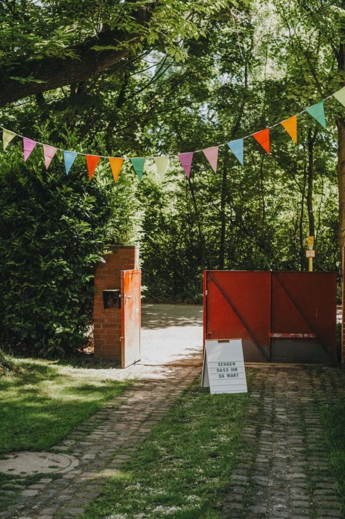 Ein mit Gras und Bäumen gesäumter Weg führt zu einem offenen roten Tor, das mit bunten Wimpeln geschmückt ist. Das Sonnenlicht dringt durch das Blätterdach - eine perfekte Szene für einen Kinderfotografen oder Werbefotografen aus einem Fotostudio Köln, um sie festzuhalten.