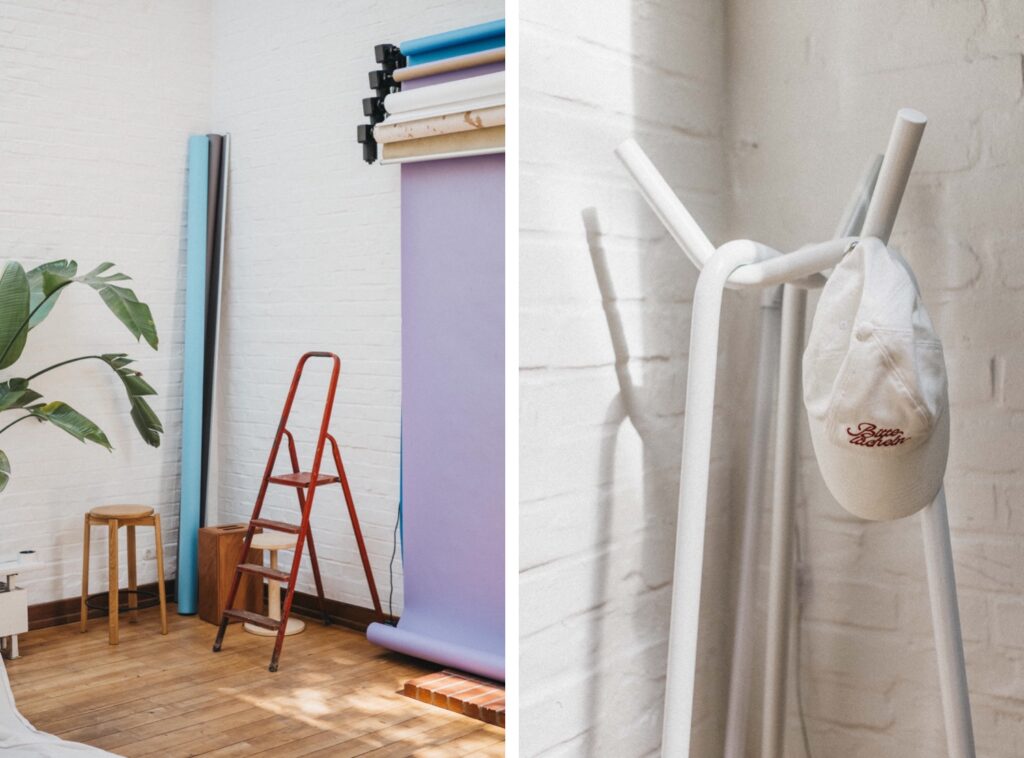 Ein helles Studio von Fotograf Köln mit einer roten Trittleiter, Holzhockern und gerollten Fotokulissen; rechts eine Nahaufnahme eines weißen Huts auf einem weißen Kleiderständer vor einer weißen Backsteinwand.