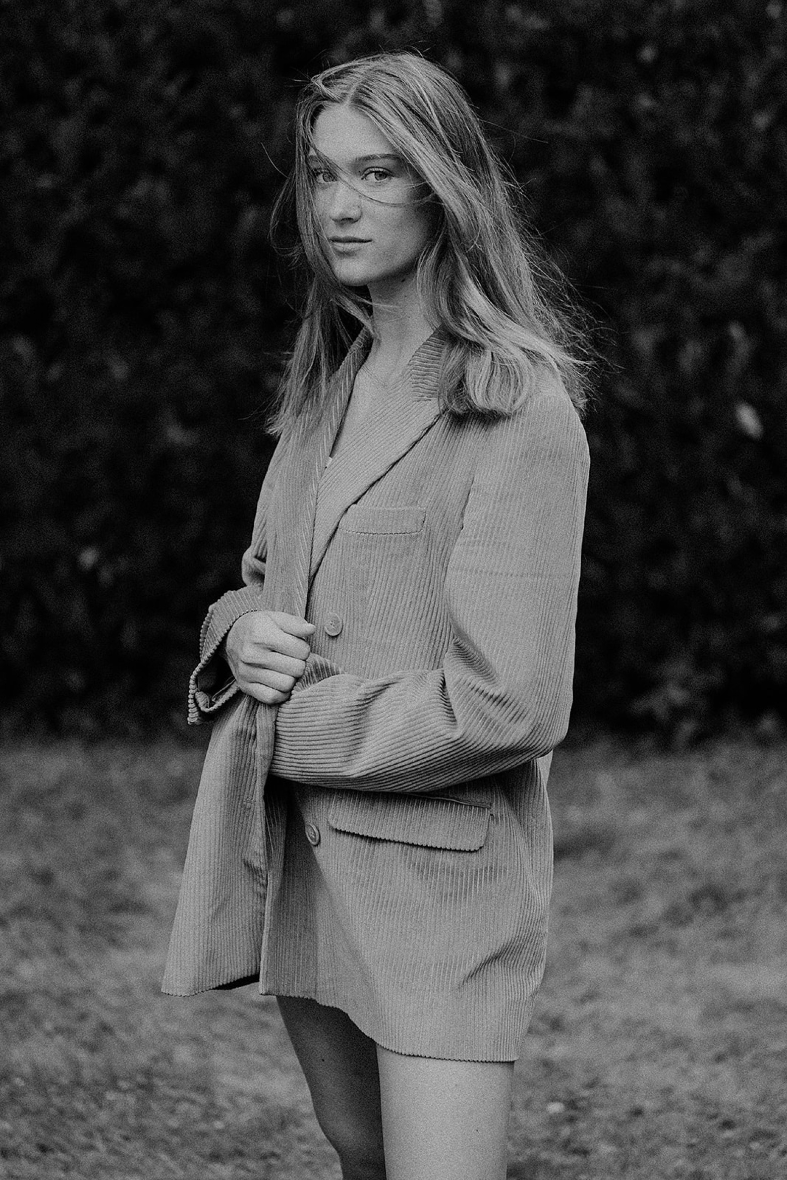 Eine junge Frau mit langen Haaren steht im Freien, gestylt in einem übergroßen Blazer. Aufgenommen von einem talentierten Werbefotografen, blickt sie mit neutralem Blick inmitten von unscharfem Laub und Gras in die Kamera. Das Foto ist in schwarz-weiß gehalten.
