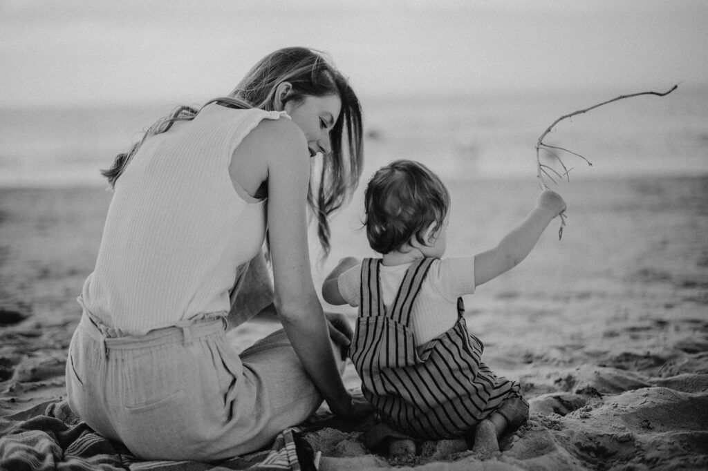 Eine Frau und ein kleines Kind sitzen bei Sonnenuntergang am Strand. Die friedliche Schwarz-Weiß-Szene, die von einem Familienfotografen aufgenommen wurde, zeigt das Kind im gestreiften Overall, das einen Stock hält, während die Frau liebevoll zuschaut.