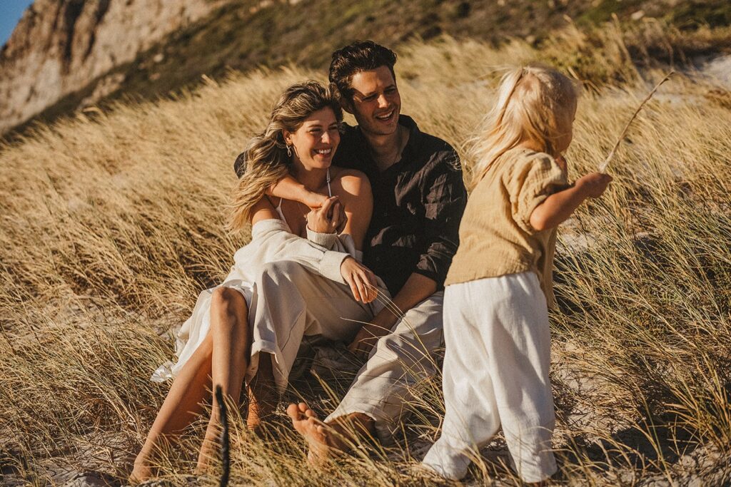 Ein lächelndes Paar sitzt zusammen im hohen Strandgras und umarmt sich, während ein kleines Kind in heller Kleidung in der Nähe spielt und einen Stock hält. Das warme Sonnenlicht, das von einem Kinderfotografen aufgenommen wurde, schafft eine entspannte, fröhliche Atmosphäre.