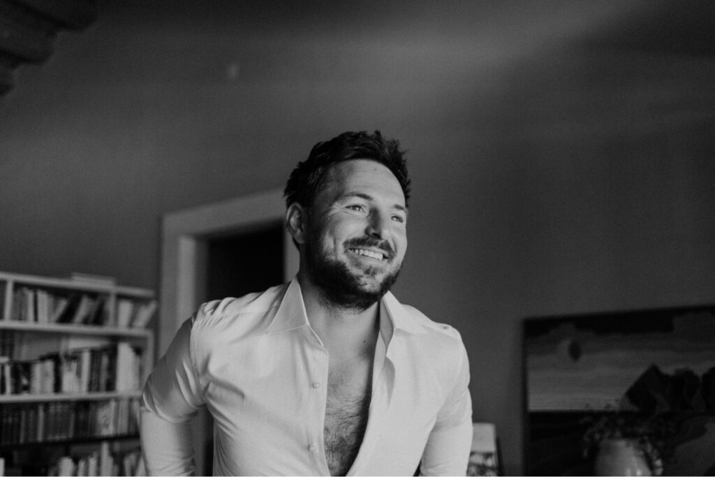 Ein Mann mit Bart und offenem Hemd lächelt, während er in einem Haus steht; im Hintergrund sind Bücherregale und ein gerahmtes Gemälde zu sehen. Das Schwarz-Weiß-Foto erinnert an die Eleganz einer klassischen sizilianischen Hochzeit.