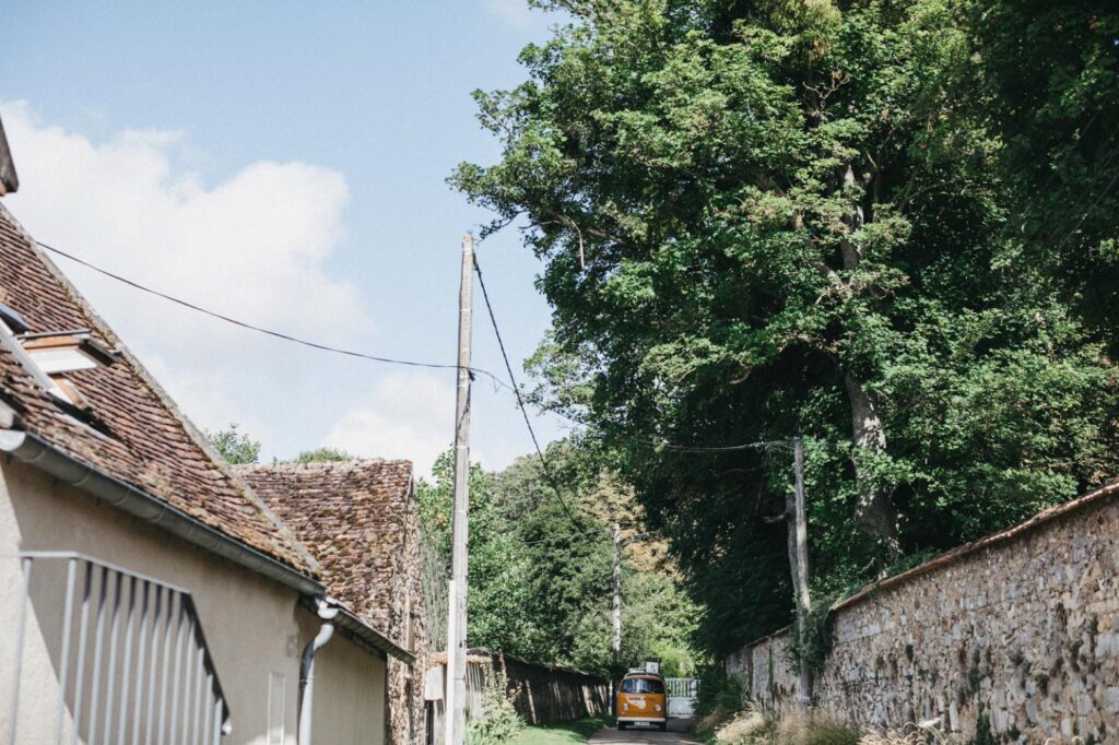 Ein gelber Lieferwagen parkt auf einer schmalen, von Steinmauern und hohen grünen Bäumen gesäumten Landstraße, aufgenommen von einem Familien Fotograf Köln, mit einem Haus auf der linken Seite und einem klaren blauen Himmel darüber.