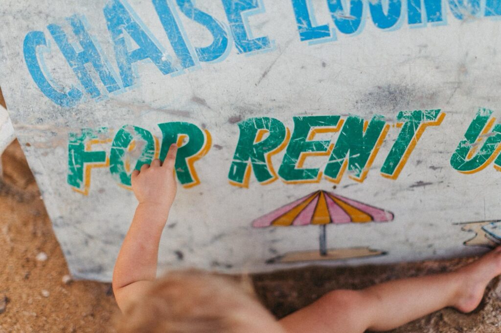 Die Hand eines Kindes berührt ein verwittertes Schild mit der Aufschrift "FOR RENT" (zu vermieten), auf dem ein Sonnenschirm abgebildet ist, umgeben von Sand - aufgenommen von einem Familienfotografen.