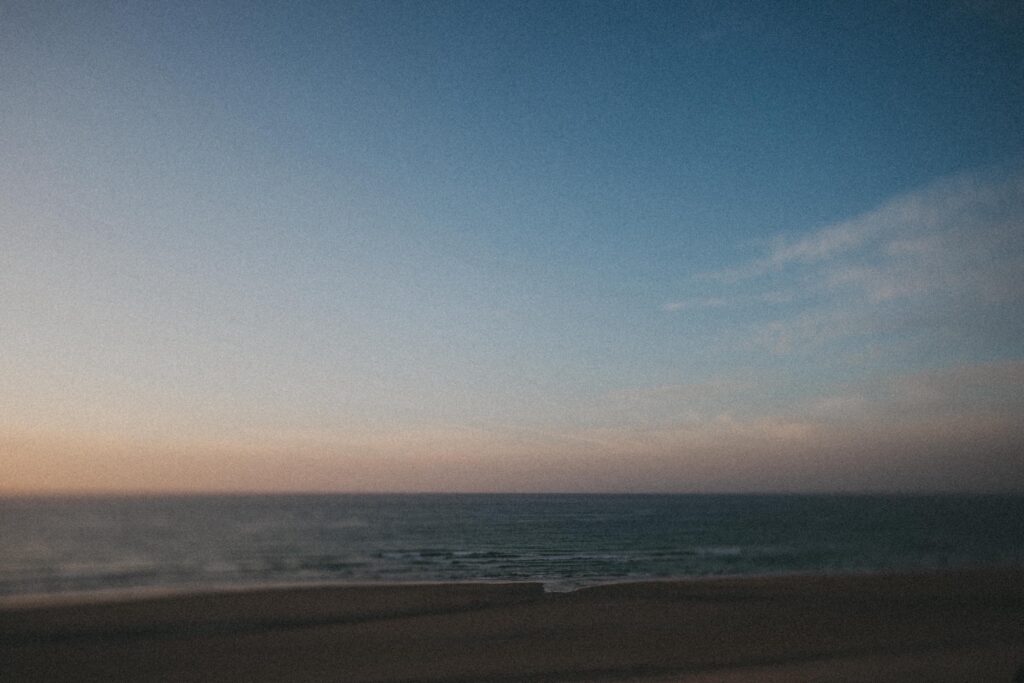 Ein unscharfes Foto eines ruhigen Ozeans mit sanften Wellen unter einem klaren Himmel, der in der Nähe des Horizonts von sanftem Orange in tiefes Blau übergeht, aufgenommen von einem Fotografen Köln, das an die Gelassenheit eines Sonnenaufgangs oder Sonnenuntergangs erinnert.