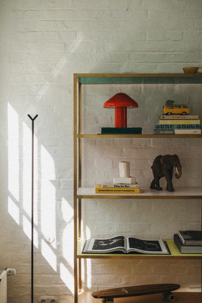 In einem sonnenbeschienenen Bücherregal vor einer weißen Backsteinwand stehen bunte Bücher, eine rote Pilzlampe, ein Spielzeugauto und eine Elefantenfigur - eingefangen von einem erfahrenen Fotografen Köln. Schatten von einem nahe gelegenen Fenster spielen über ein aufgeschlagenes Buch und ein Skateboard im unteren Regal.